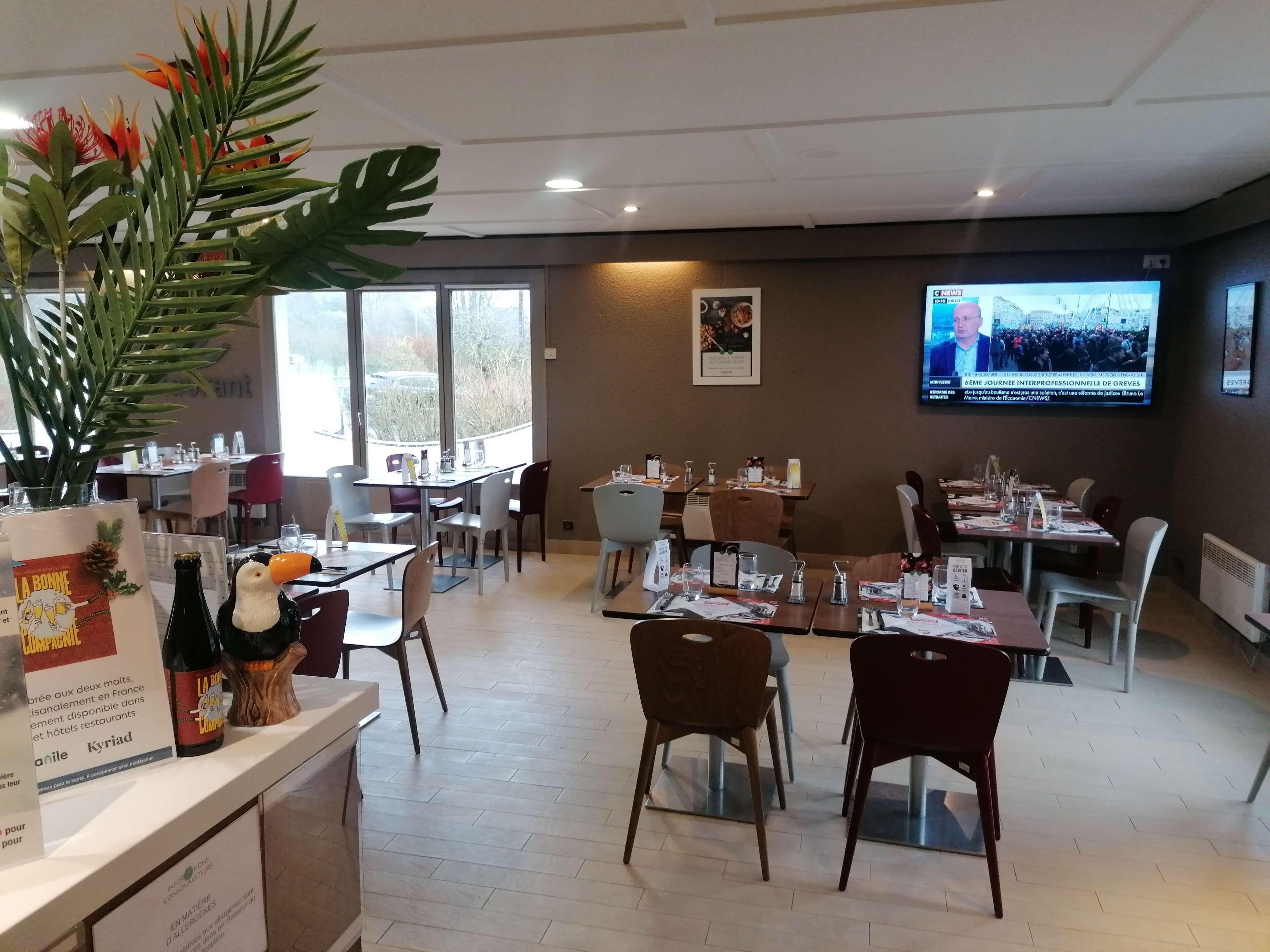 A restaurant with multiple tables and chairs, a large screen displaying news, and a view of the outdoors through large windows.