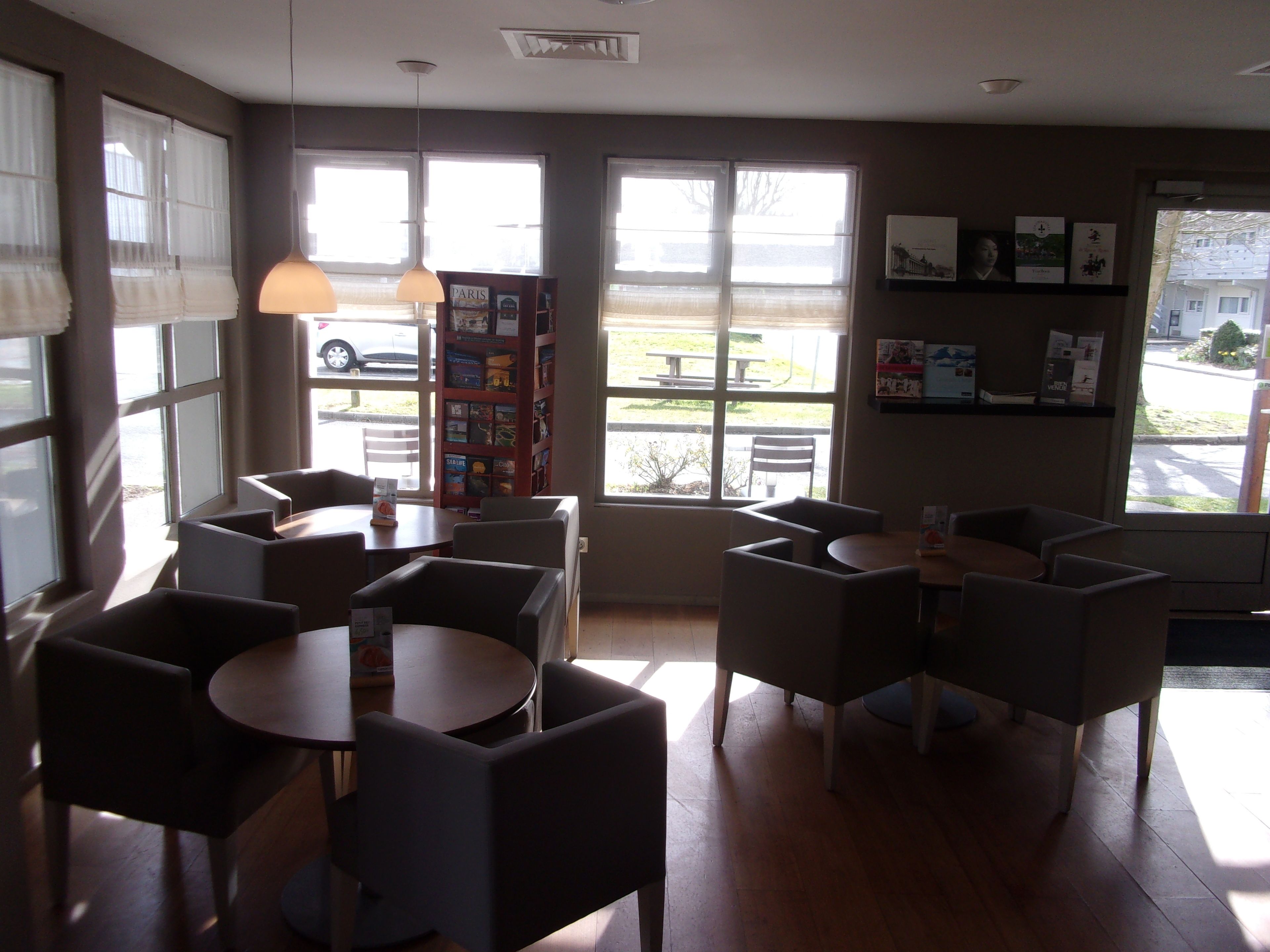 A cafe with multiple tables and chairs, large windows with blinds, and a bookshelf with books and magazines.