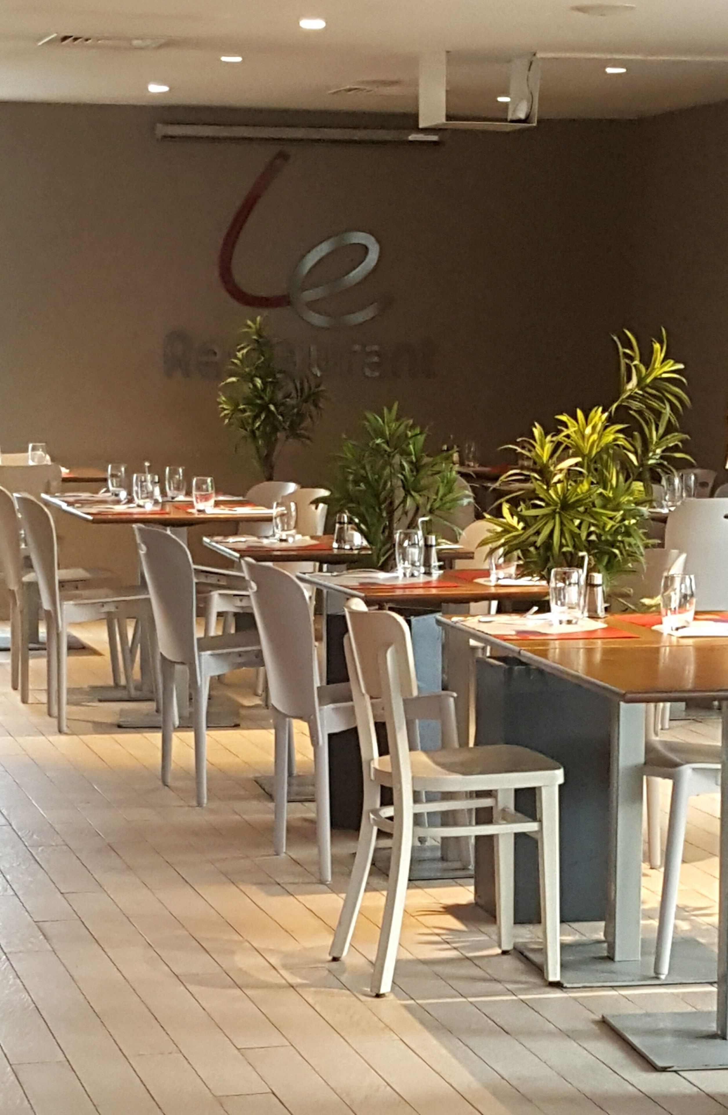Ein Restaurantinneres mit mehreren Tischen, Stühlen, Kübelpflanzen und einer Wand mit einem Logo.