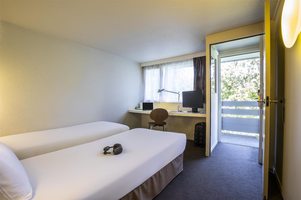 Une chambre d’hôtel avec deux lits simples, un bureau avec un ordinateur, une chaise et une vue sur l’extérieur par une porte.