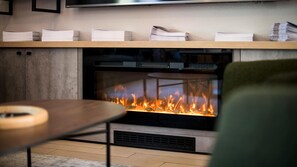 A modern fireplace with a visible fire, a wooden shelf above it, and a table in the foreground.