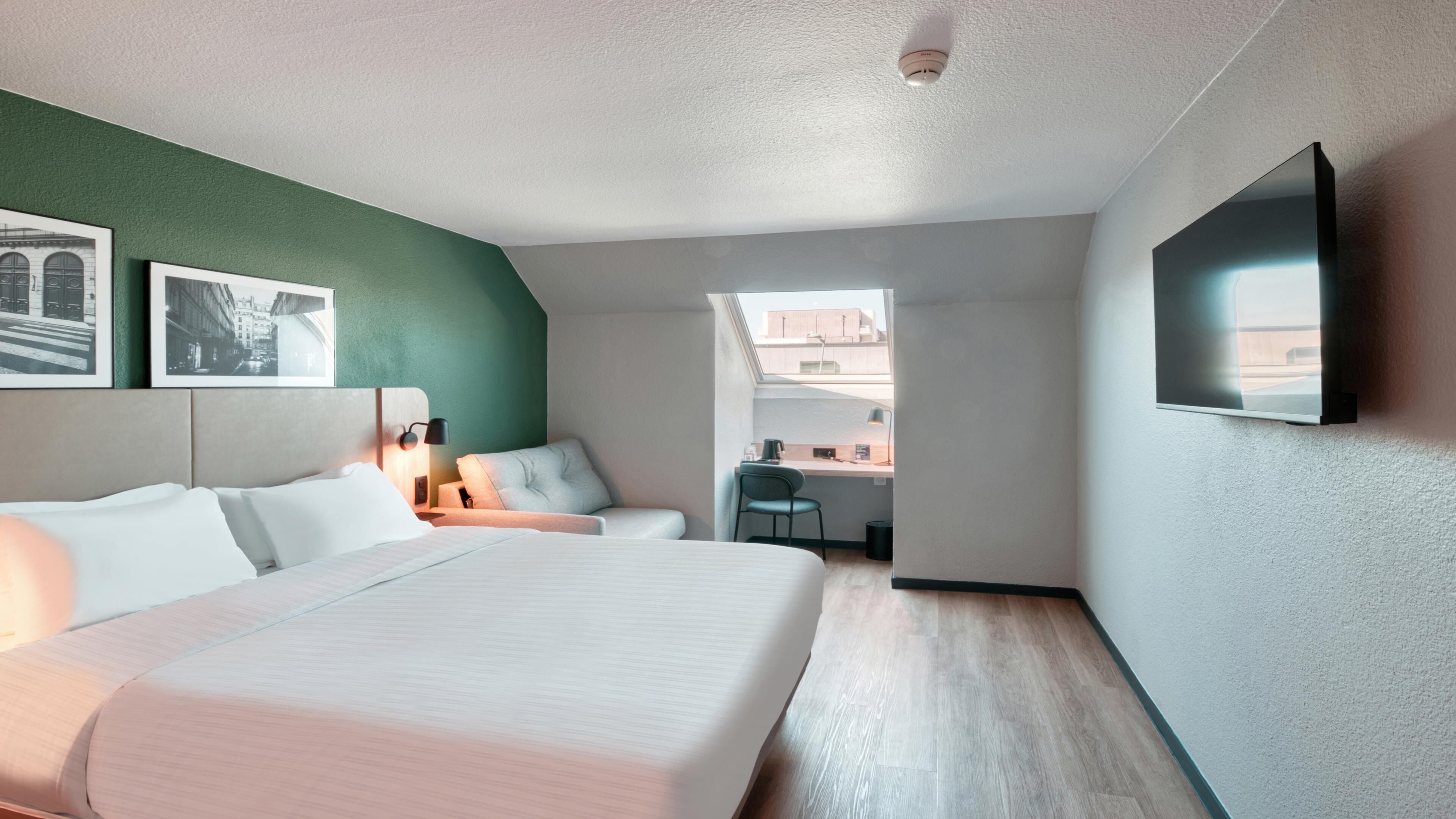 Una habitación de hotel moderna con una cama grande, un escritorio y una silla. Hay dos cuadros en la pared y un televisor de pantalla plana montado allí.