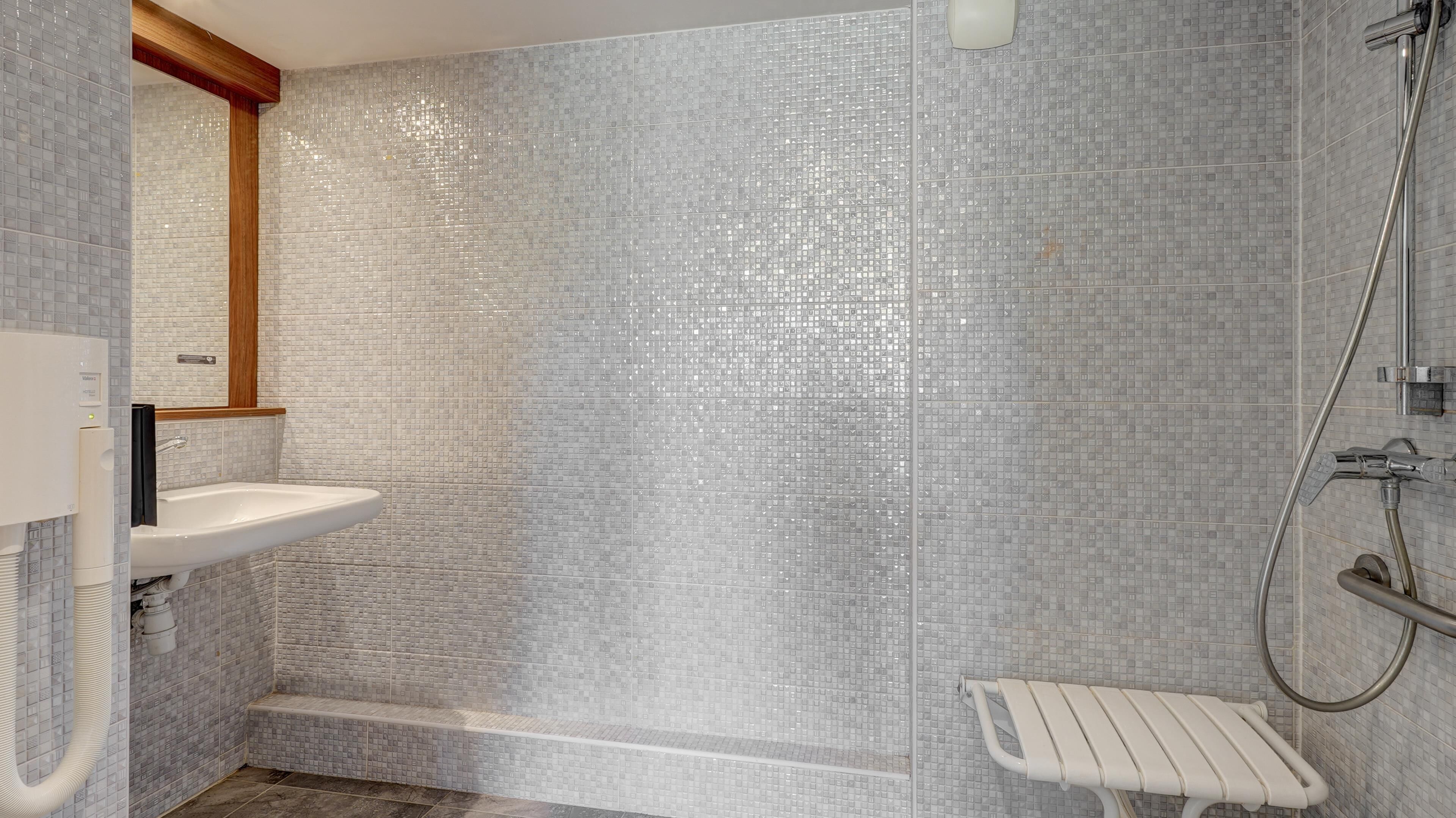 Un baño con lavamanos blanco, ducha con cabezal de ducha de mano y un banco.
