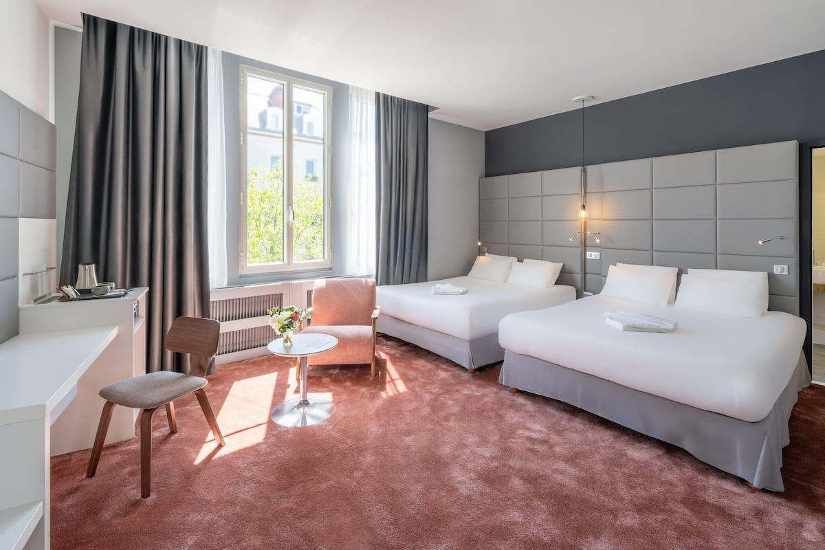 Quarto de hotel com duas camas, uma escrivaninha, uma cadeira, uma mesinha de centro com um vaso, e uma janela grande com cortinas.