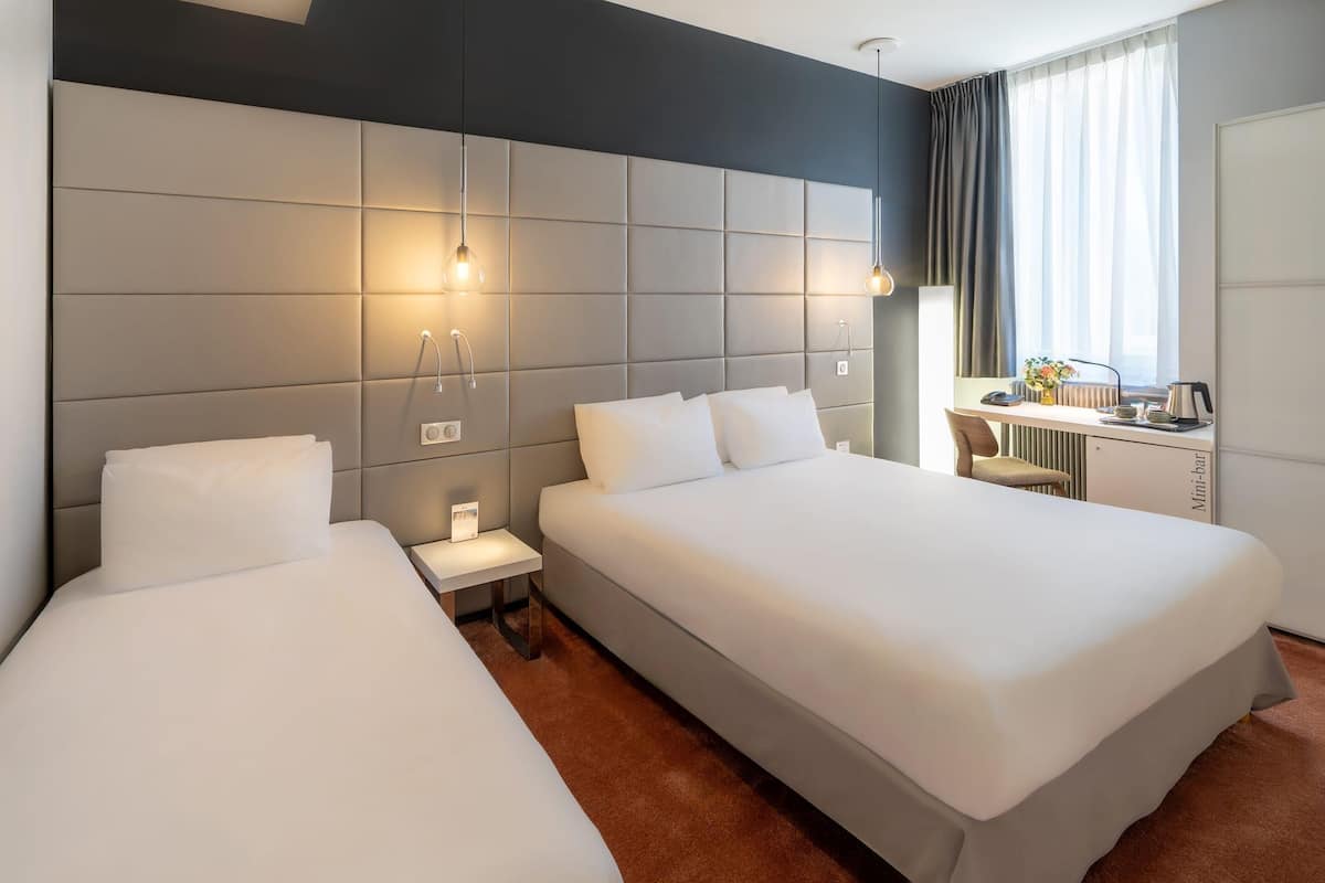 Quarto de hotel com duas camas, uma escrivaninha, uma cadeira, frigobar e janela com cortinas.