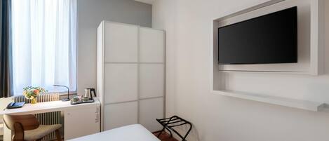 Habitación de hotel con cama, escritorio con silla, radiador, televisor montado en la pared y ventana con cortinas.