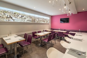 Un área de comedor moderna con mesas y sillas, un ventanal con vista a la ciudad y una pared rosa con un televisor.