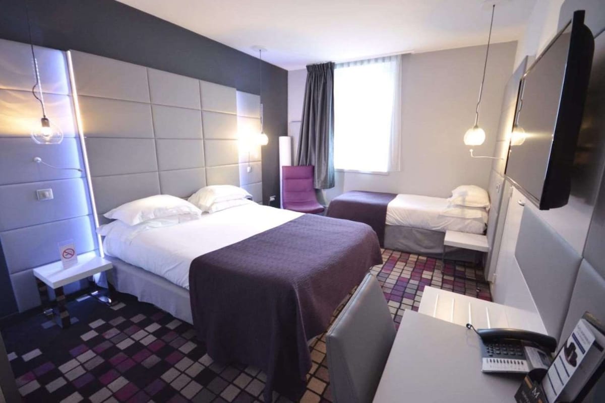 A hotel room with a bed, a desk with a phone, a chair, a small table, and a window with curtains.