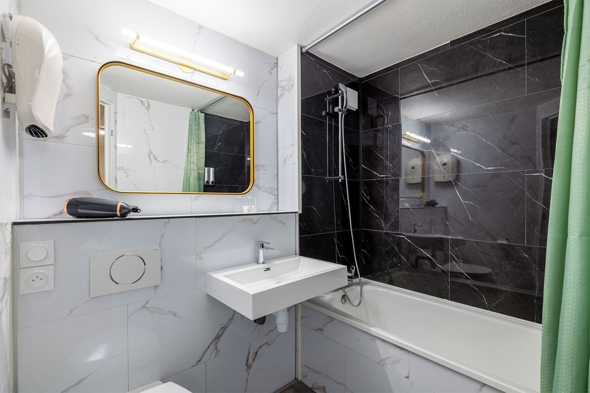 Un baño moderno con un lavamanos blanco, un espejo con marco dorado, un secador de pelo de mano y una ducha con baldosas de mármol negro.