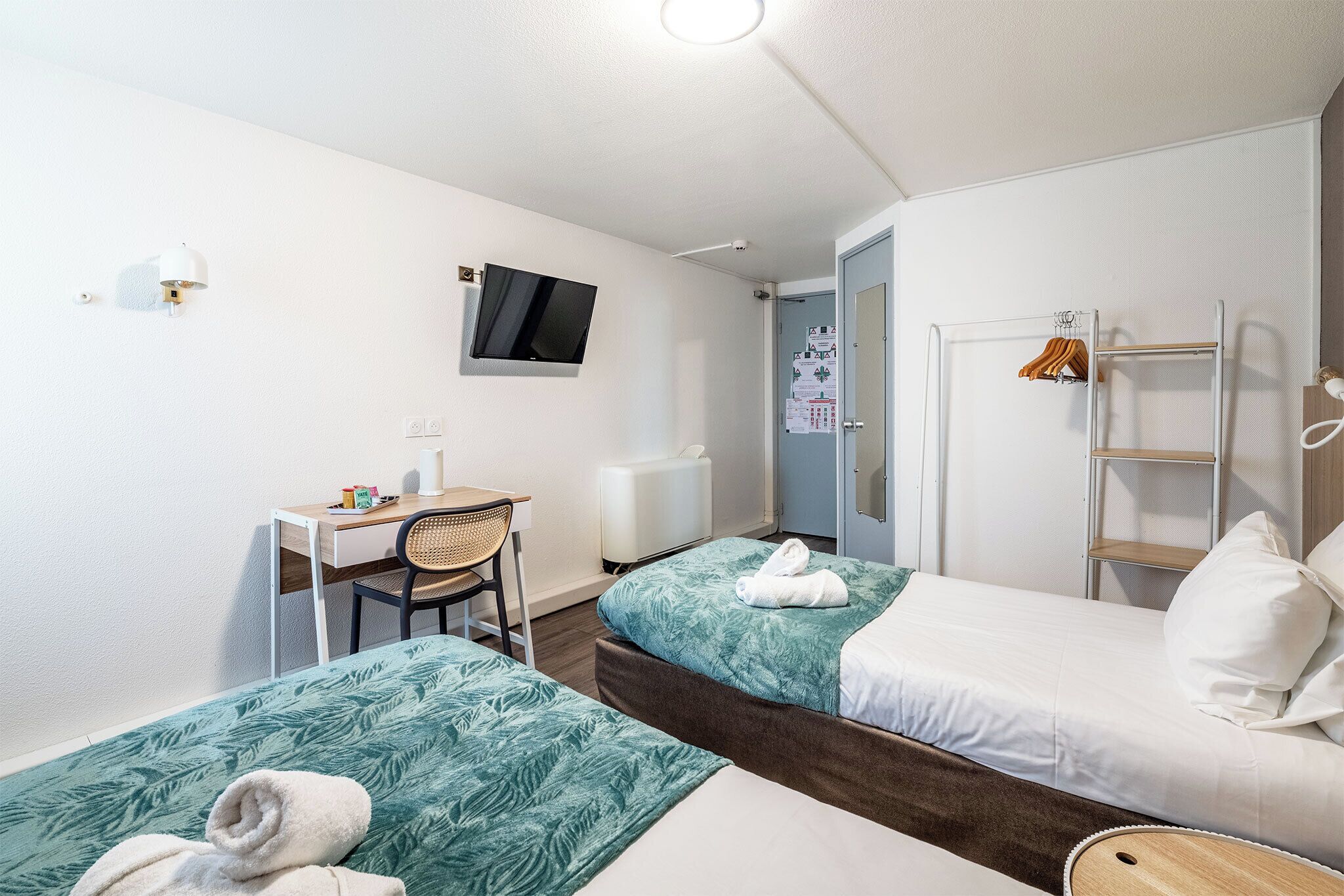 A small hotel room with two beds, a desk, a chair, a TV, and a door leading to a bathroom.