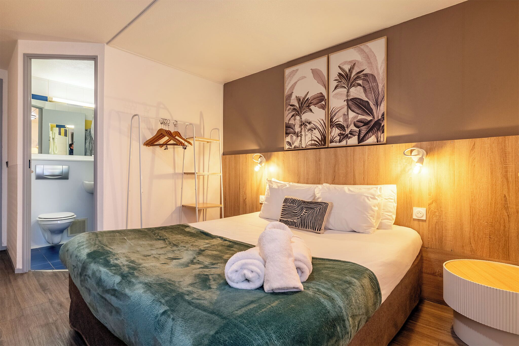 Habitación de hotel con cama, mesita de noche, luz fijada a la pared y baño visible a través de una puerta abierta.