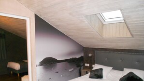 Une chambre à coucher comprenant un lit, une table de chevet, un tabouret, une fresque murale et une lucarne.