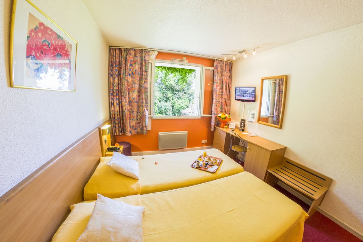 A hotel room with two beds, a window with curtains, a desk with a chair, a radiator, and a television on the wall.