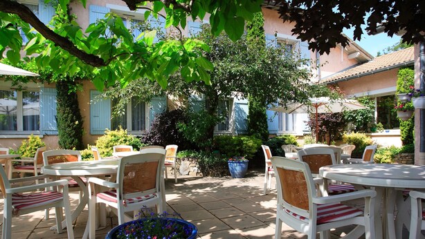 Une terrasse extérieure avec des tables et des chaises blanches, entourée d’une végétation luxuriante et de plantes en pot.