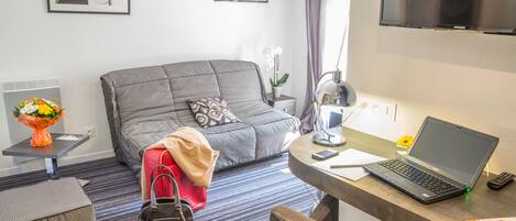 A modern hotel room with a grey sofa, a small side table with a vase of flowers, a laptop on a desk, and a flat-screen TV on the wall.
