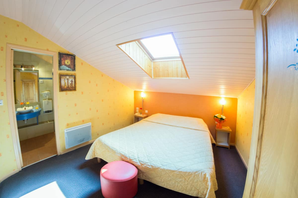 A bedroom with a bed, a skylight, a nightstand with a lamp, and a bathroom visible through an open door.