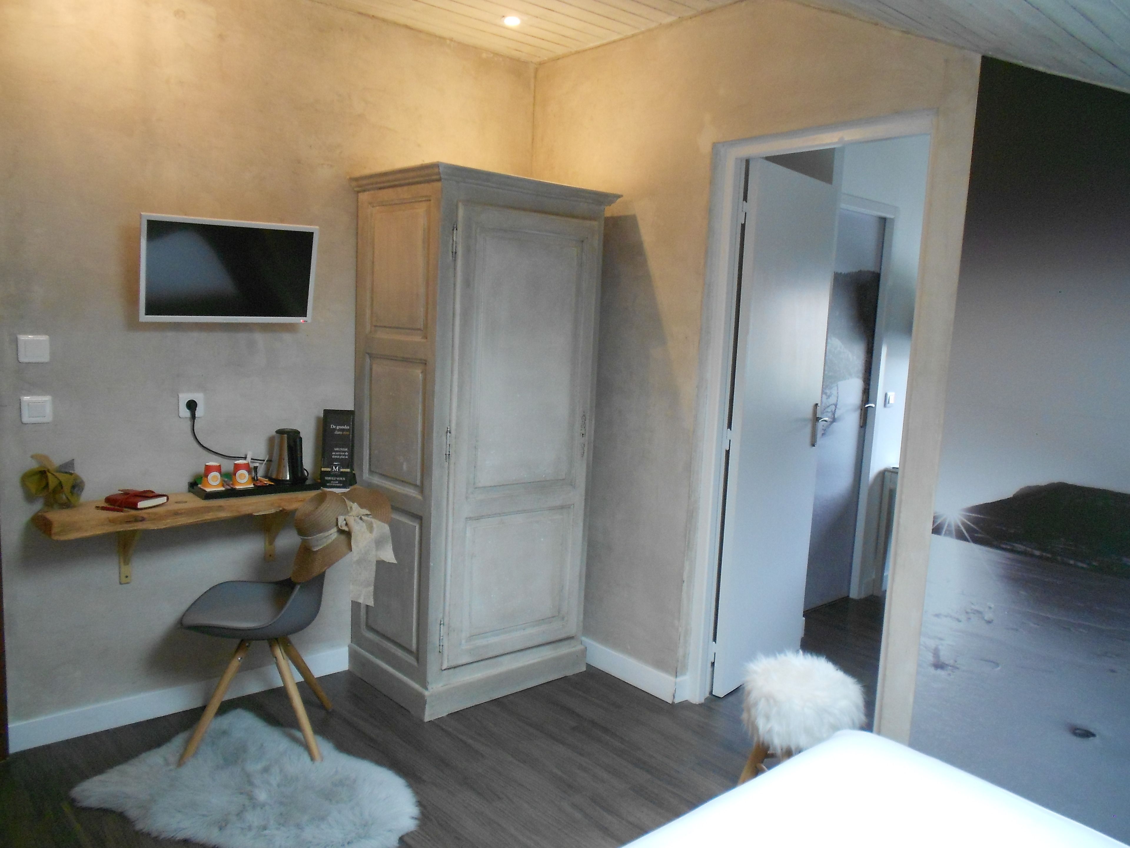 A bedroom with a bed, a desk with a chair, a wardrobe, a television on the wall, and a door leading to another room.