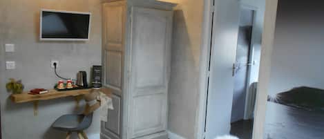 Un dormitorio con una cama, un escritorio con silla, un armario, un televisor en la pared y una puerta que da a otra habitación.