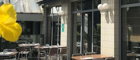 Un área de comedor al aire libre con mesas y sillas, ventanas amplias y un letrero de salida verde.