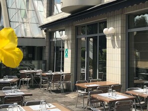 Un espace extérieur aménagé pour les repas, avec des tables et des chaises, de grandes fenêtres et un panneau de sortie vert.