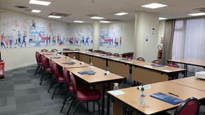 Une salle de conférence avec de longues tables, des chaises rouges, des plaques nominatives et un extincteur.