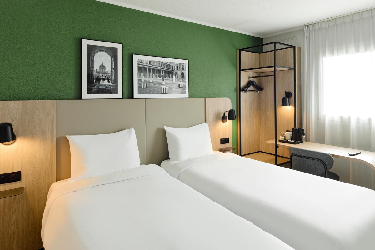 Ein Hotelzimmer mit zwei Betten, einem Schreibtisch, einem Stuhl, einem Kleiderhaken und zwei gerahmten Bildern an der Wand.