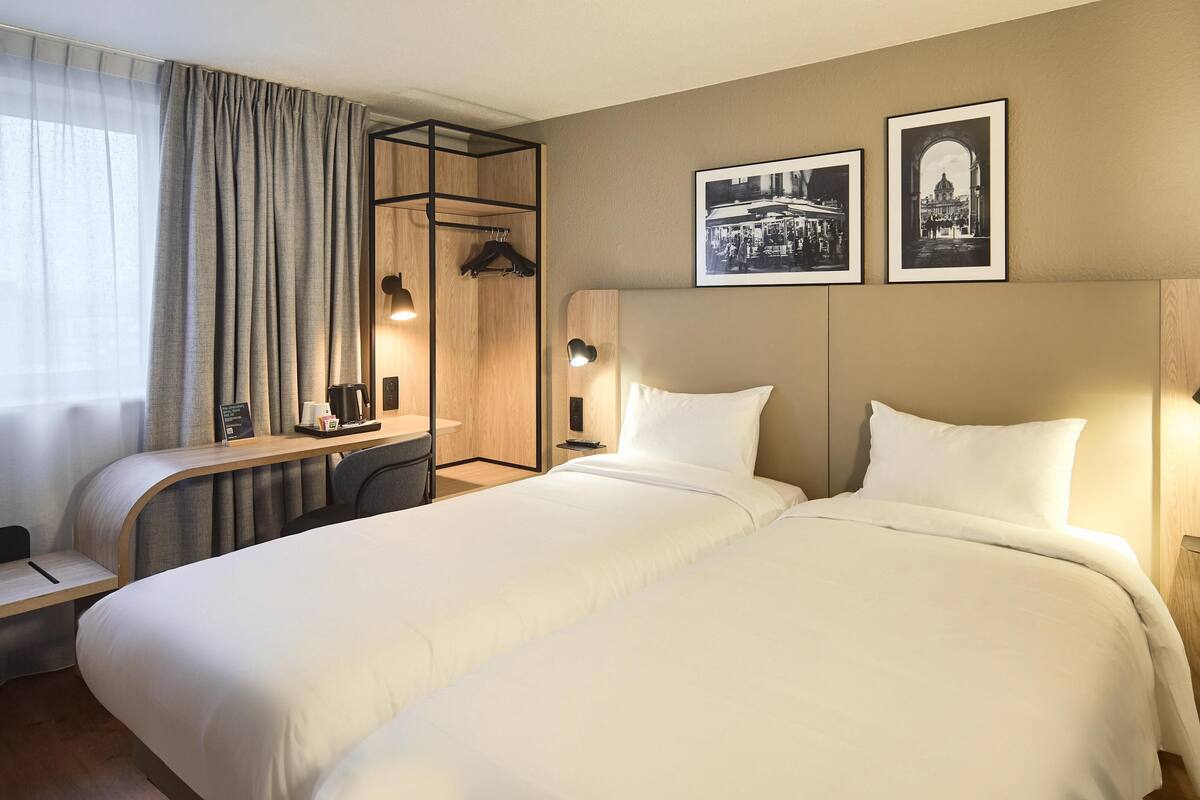 Une chambre d’hôtel avec un lit, un bureau, une chaise, un porte-manteau et deux tableaux encadrés au mur.
