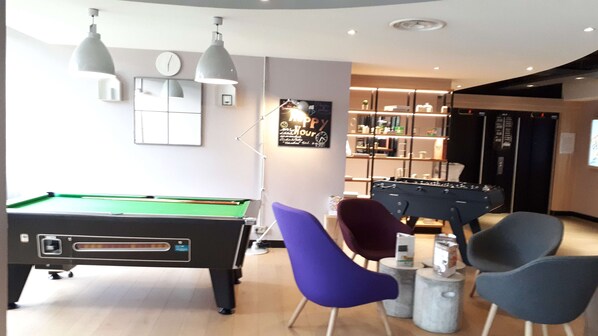 A pool table, two chairs, a foosball table, and a bar area with shelves and a clock on the wall.