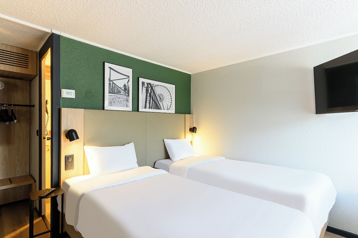 A hotel room with two beds, a wooden wardrobe, a television mounted on the wall, and two framed pictures on the wall.
