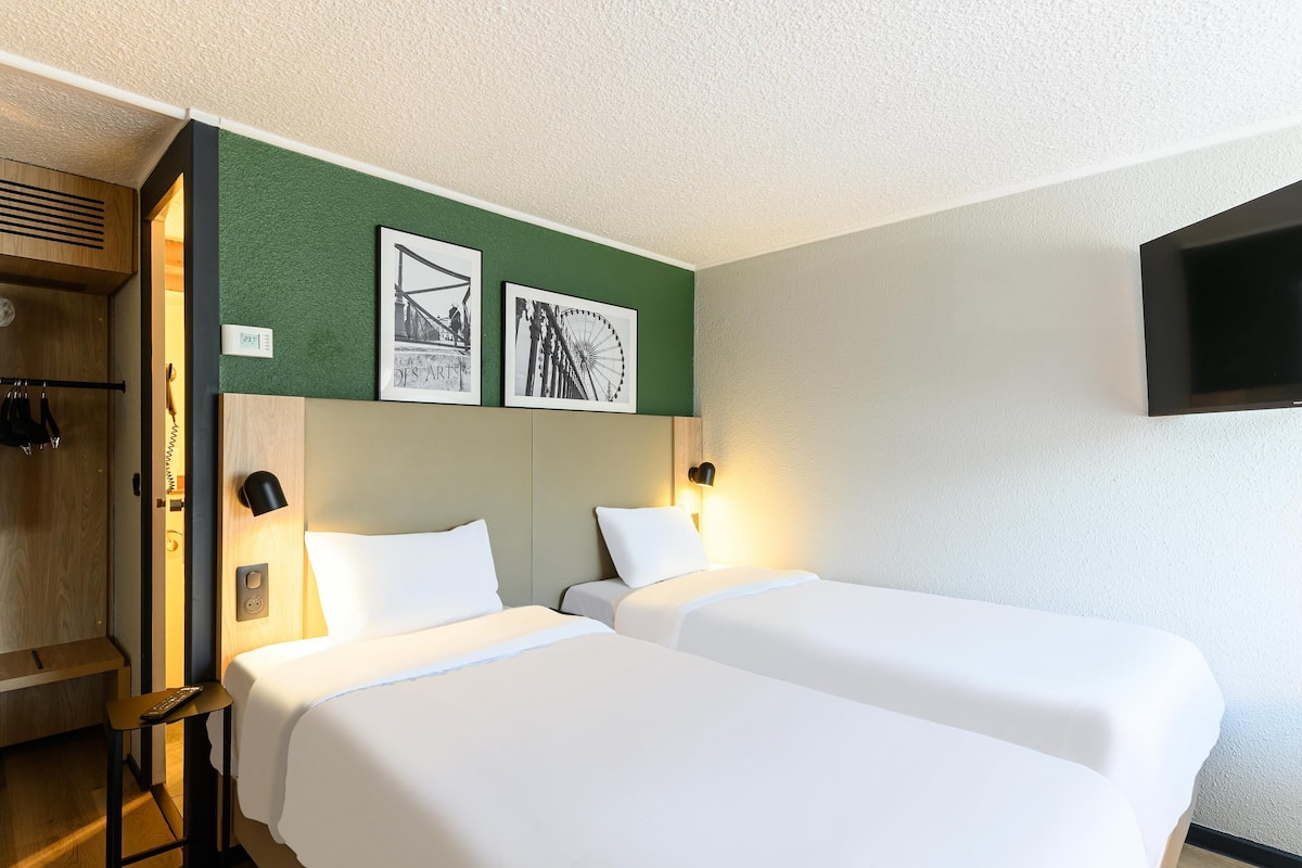 A hotel room with two beds, a wooden wardrobe, a television mounted on the wall, and two framed pictures on the wall.