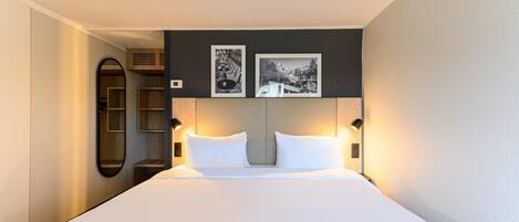 Une chambre d’hôtel avec un grand lit, deux lampes de chevet, un miroir et deux tableaux encadrés au mur.