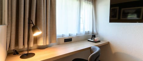Une chambre d’hôtel avec un bureau en bois, une lampe, une chaise, une fenêtre avec des rideaux et un cadre photo fixé au mur.