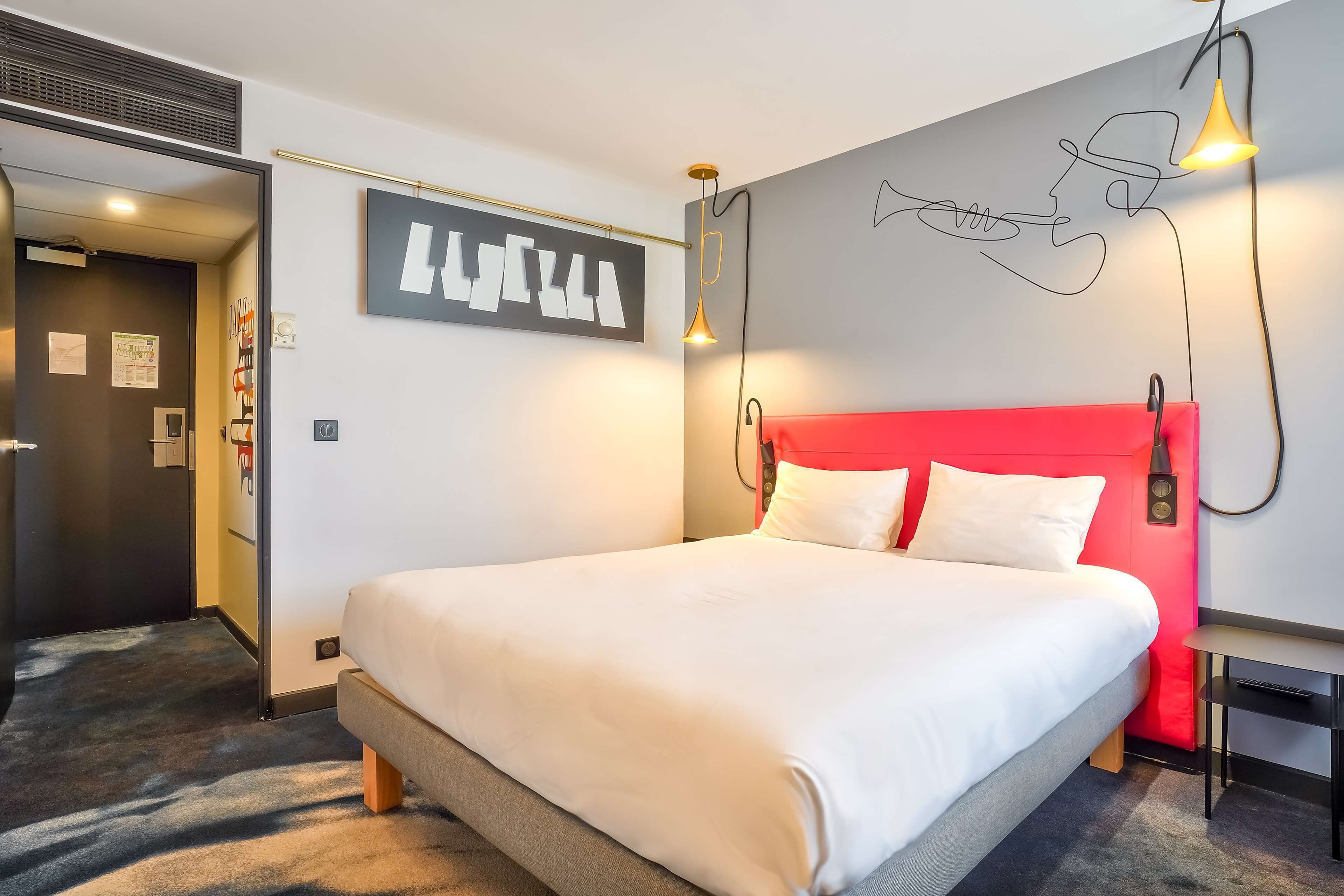 A hotel room with a large bed, a red headboard, a desk, and a wall-mounted light fixture.