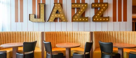 Interior de restaurante con temática de jazz, un gran letrero iluminado que dice 'JAZZ', asientos acolchados y mesas redondas.