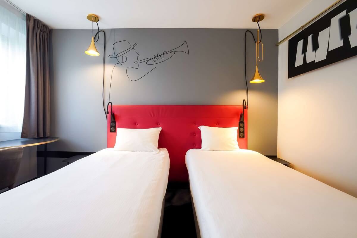 Hotellihuone, jossa on kaksi sänkyä, punainen pääty ja seinällä oleva taideteos, jossa on kuvattuna trumpetti.