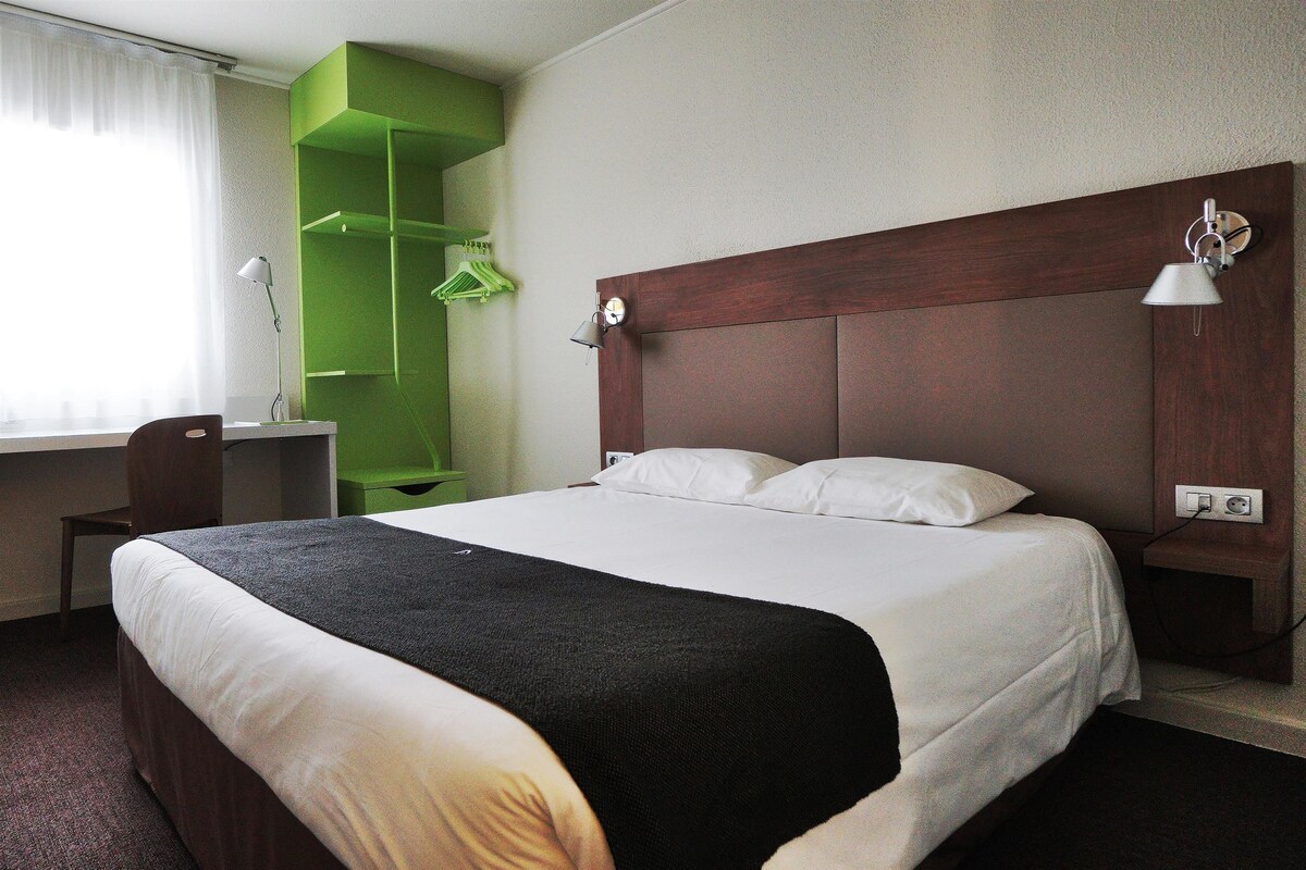 A hotel room with a large bed, a desk, a chair, a green shelving unit, and a wall-mounted lamp.