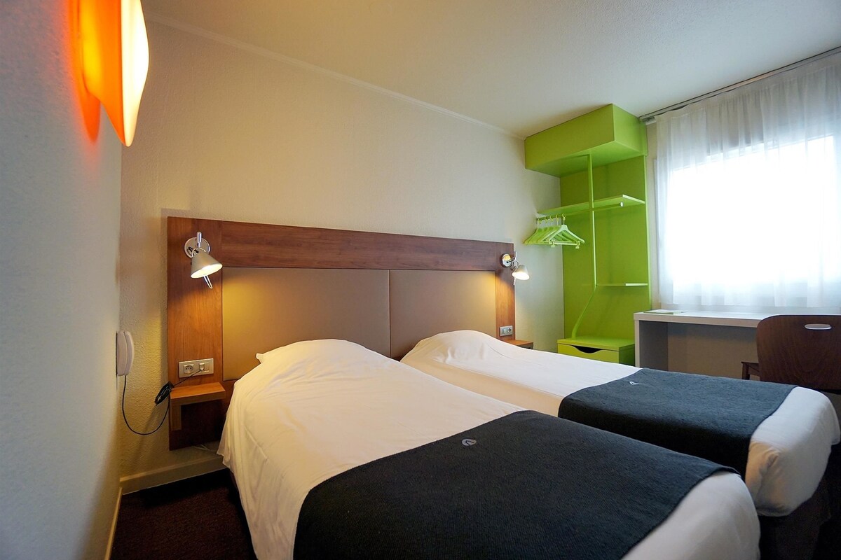 A hotel room with two beds, a desk, a chair, a green wardrobe, and a window with curtains.
