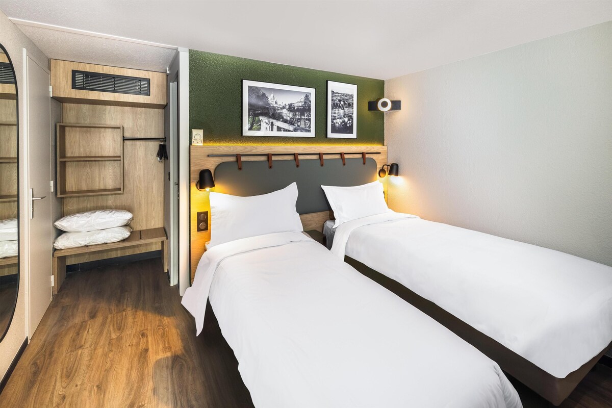 Ein Hotelzimmer mit zwei Einzelbetten, einem Holzschrank, einer Sitzbank mit Handtüchern und zwei gerahmten Bildern an der Wand.