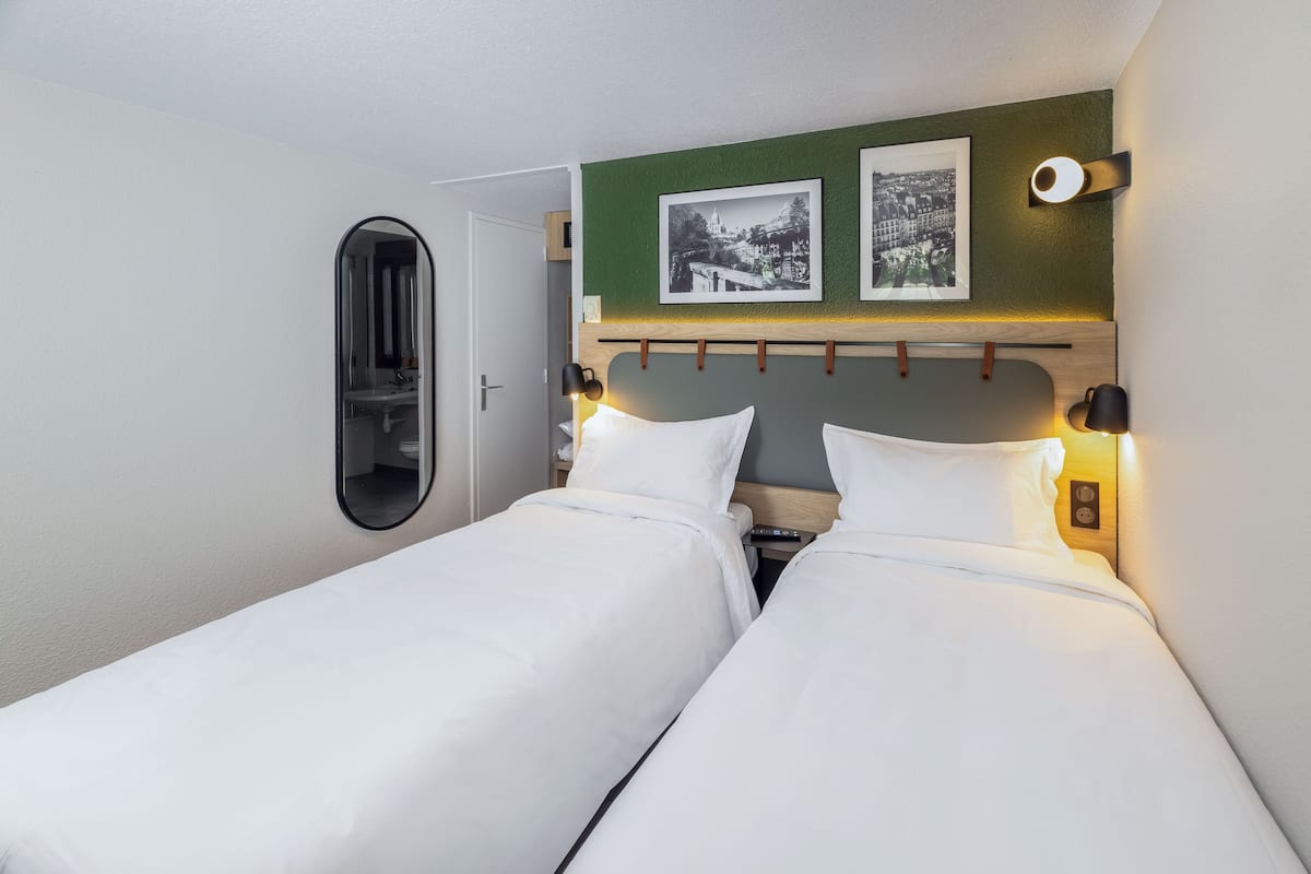 A hotel room with two single beds, a mirror, a green accent wall, and two framed pictures.