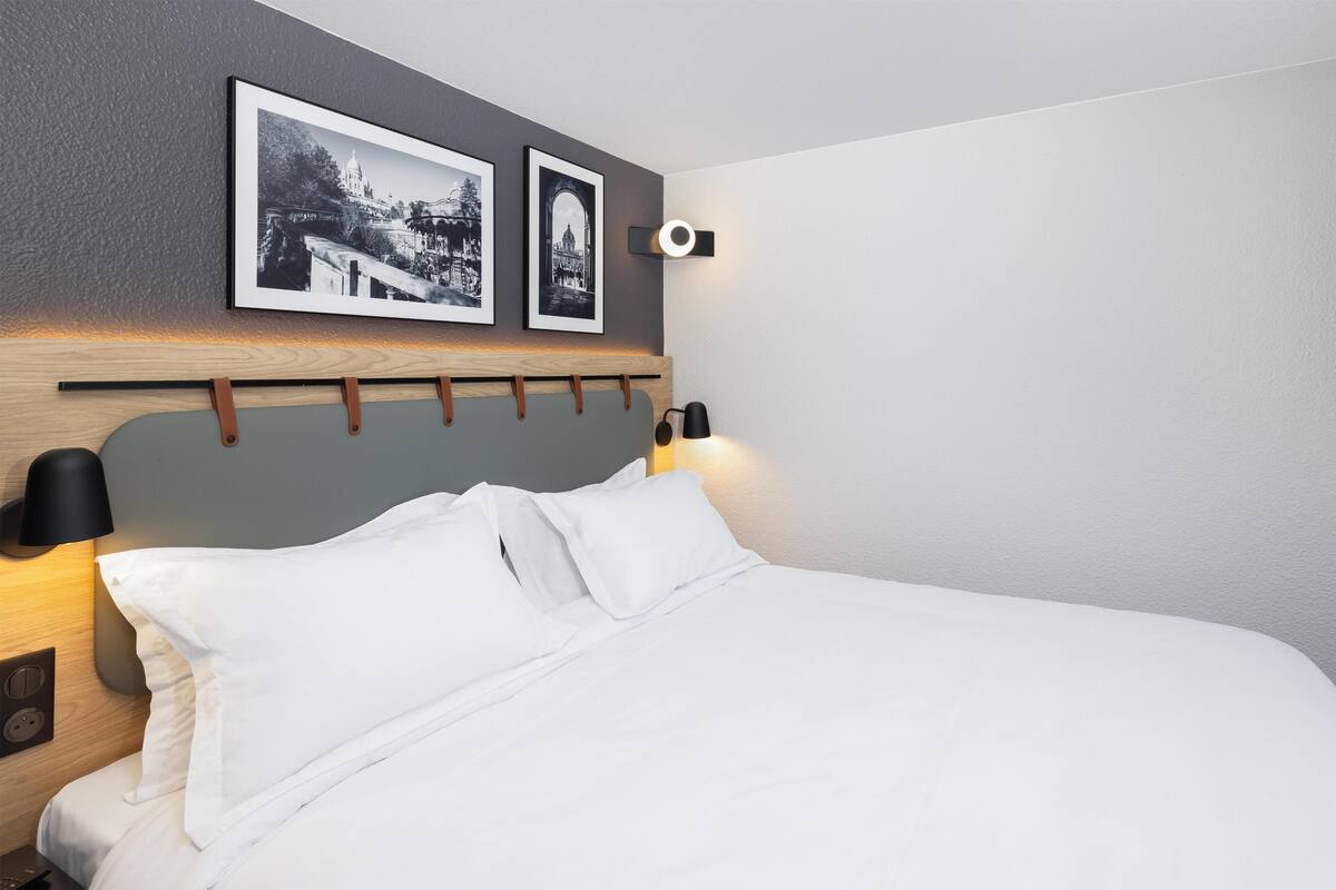 A neatly made bed with white linens, a headboard with a wooden panel, and two black wall sconces.