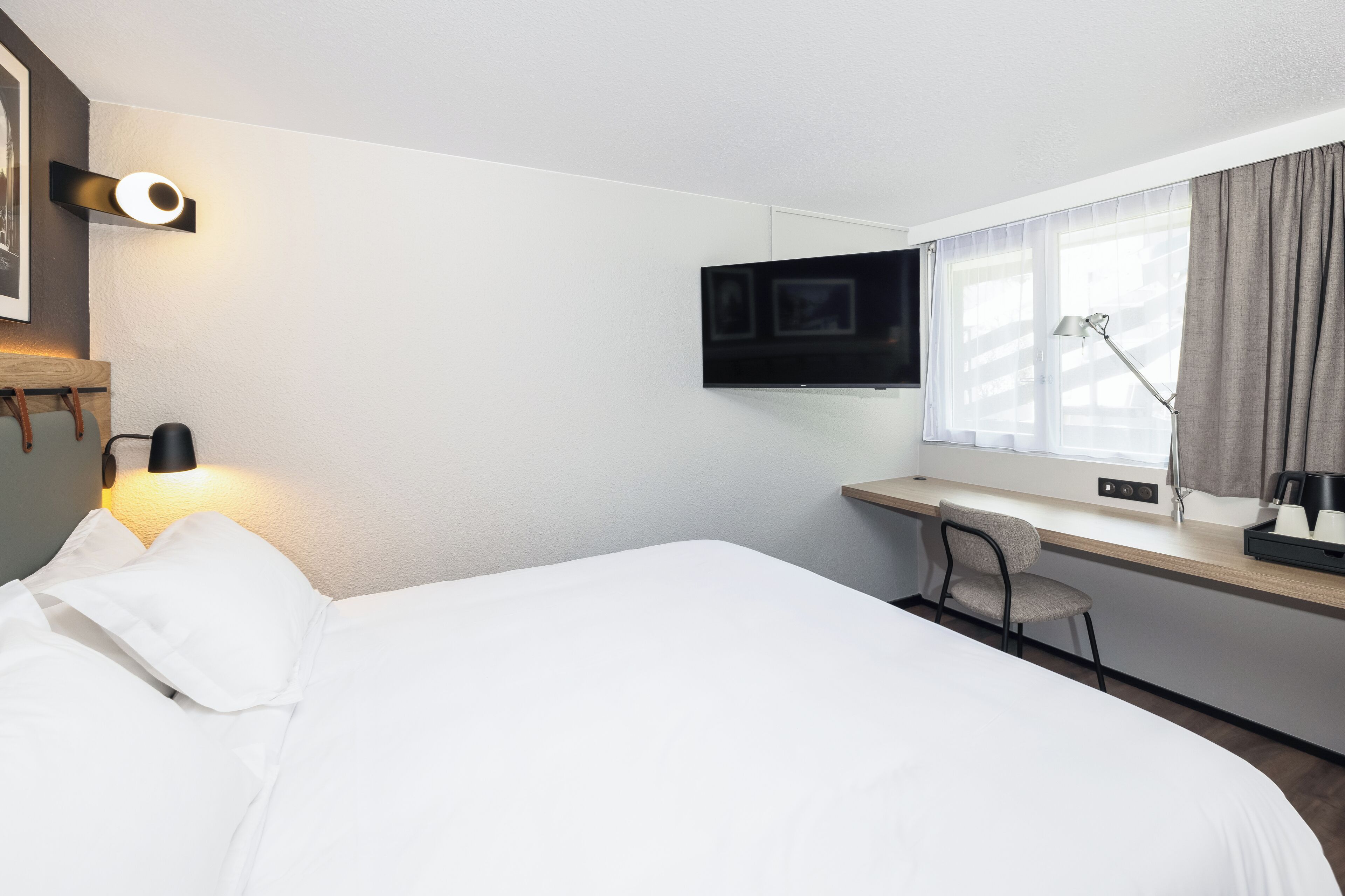 A hotel room with a bed, a desk, a chair, a TV mounted on the wall, and a window with curtains.