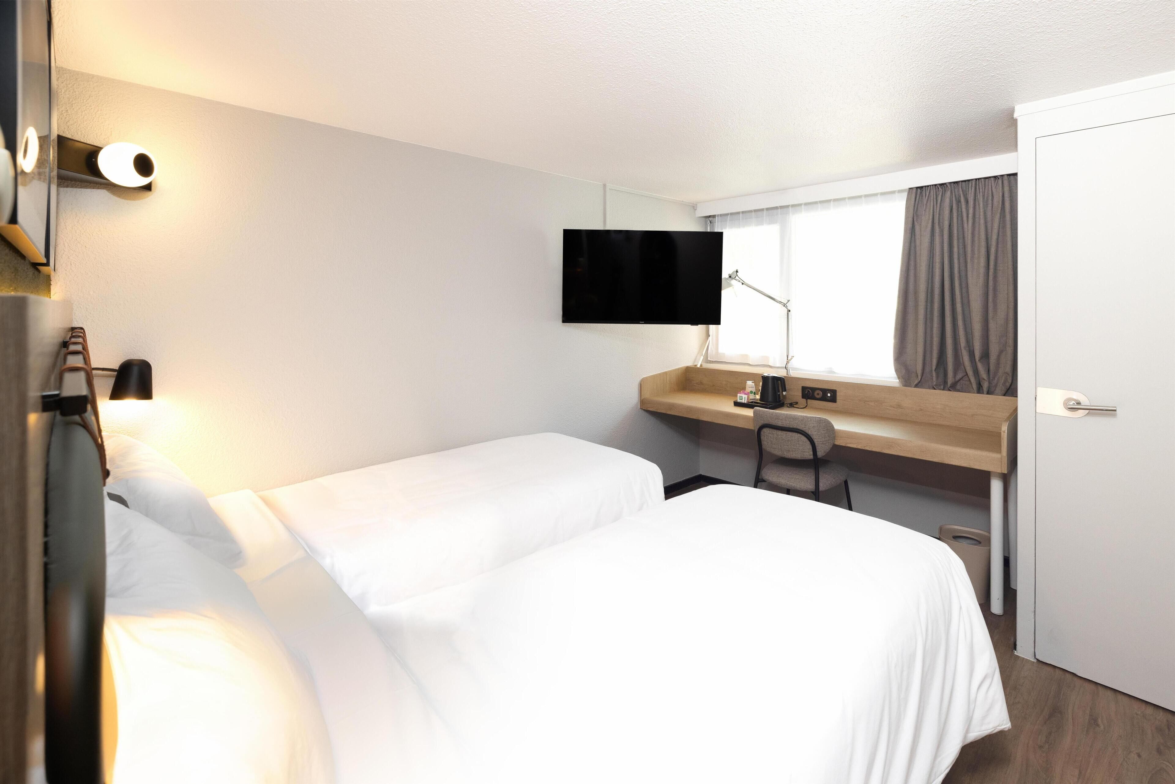 Une chambre d’hôtel avec deux lits simples, un bureau avec une chaise, un téléviseur fixé au mur et une fenêtre avec des rideaux.