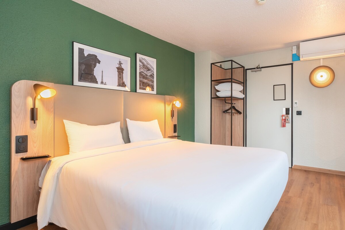 A hotel room with a bed, two wall-mounted lights, a wardrobe, a door with a handle, and a wall-mounted air conditioning unit.