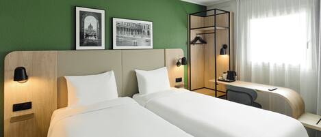 A hotel room with two beds, a desk, a chair, a coat rack, and two framed pictures on the wall.