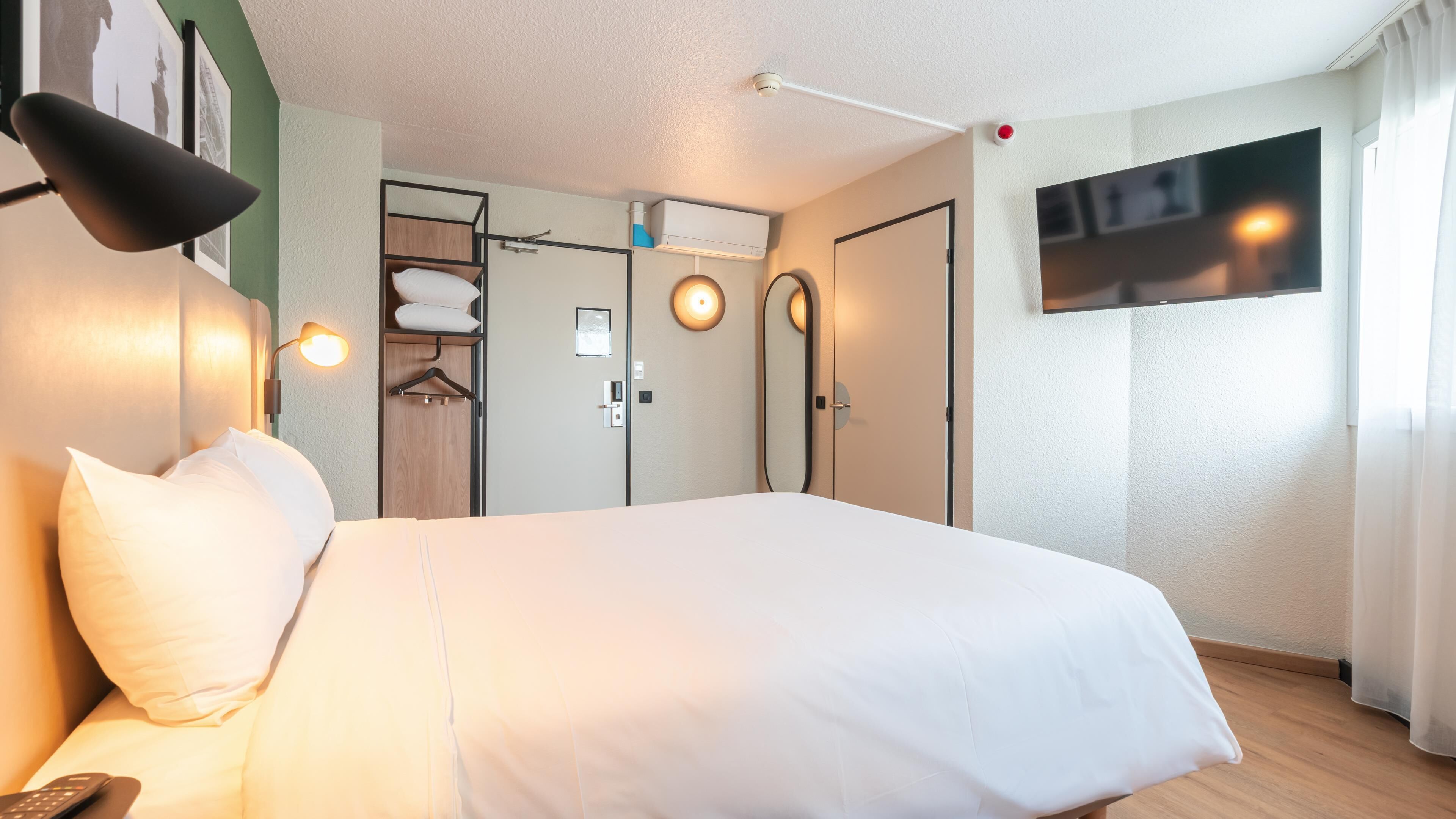 A hotel room with a bed, a wardrobe, a mirror, a TV, and a ceiling-mounted light fixture.