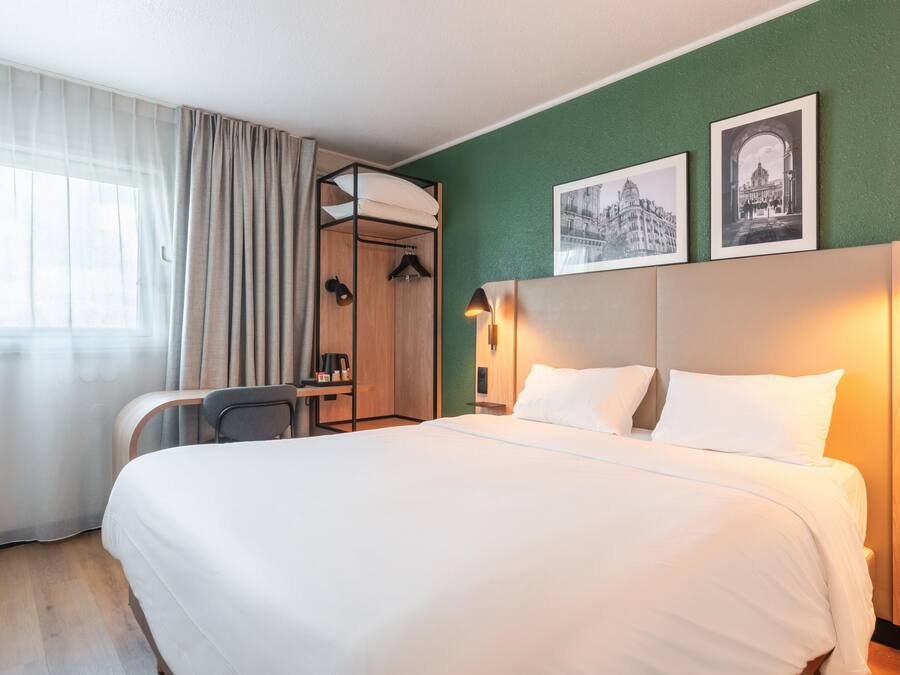 A hotel room with a bed, a desk, a chair, a wardrobe, and two framed pictures on the wall.