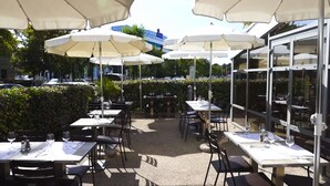 An outdoor dining area with tables and chairs under white umbrellas, surrounded by greenery and a glass building.