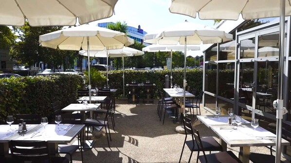 An outdoor dining area with tables and chairs under white umbrellas, surrounded by greenery and a glass building. - Campanile PRIME - Paris Gennevilliers Barbanniers (Gennevilliers)