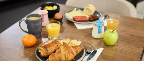Una mesa de desayuno con cruasanes, jugo de naranja, una manzana verde, un hot dog y un plato de huevos.
