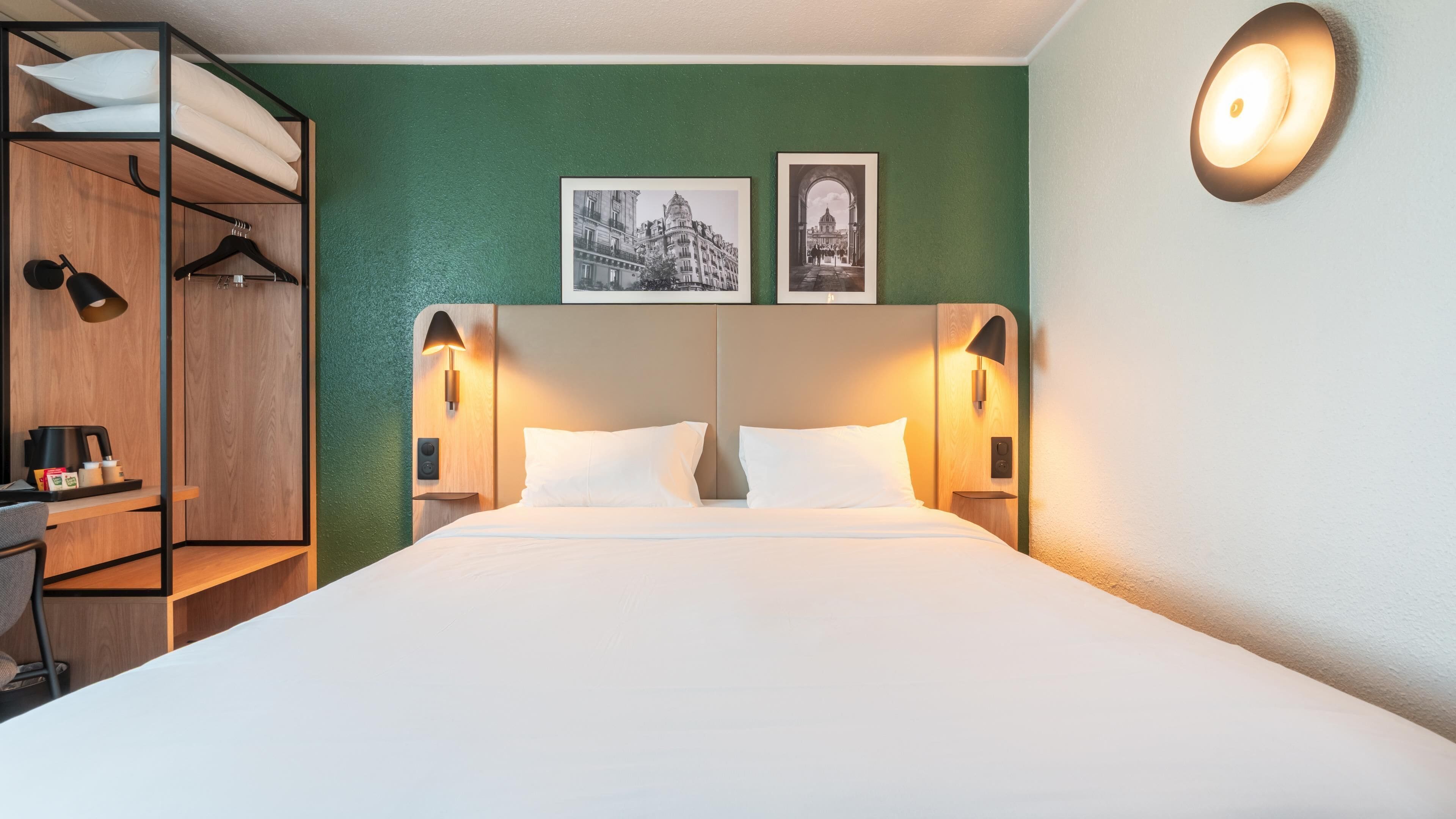 A hotel room with a bed, two wall-mounted lamps, a desk with a coffee maker, and a wardrobe.